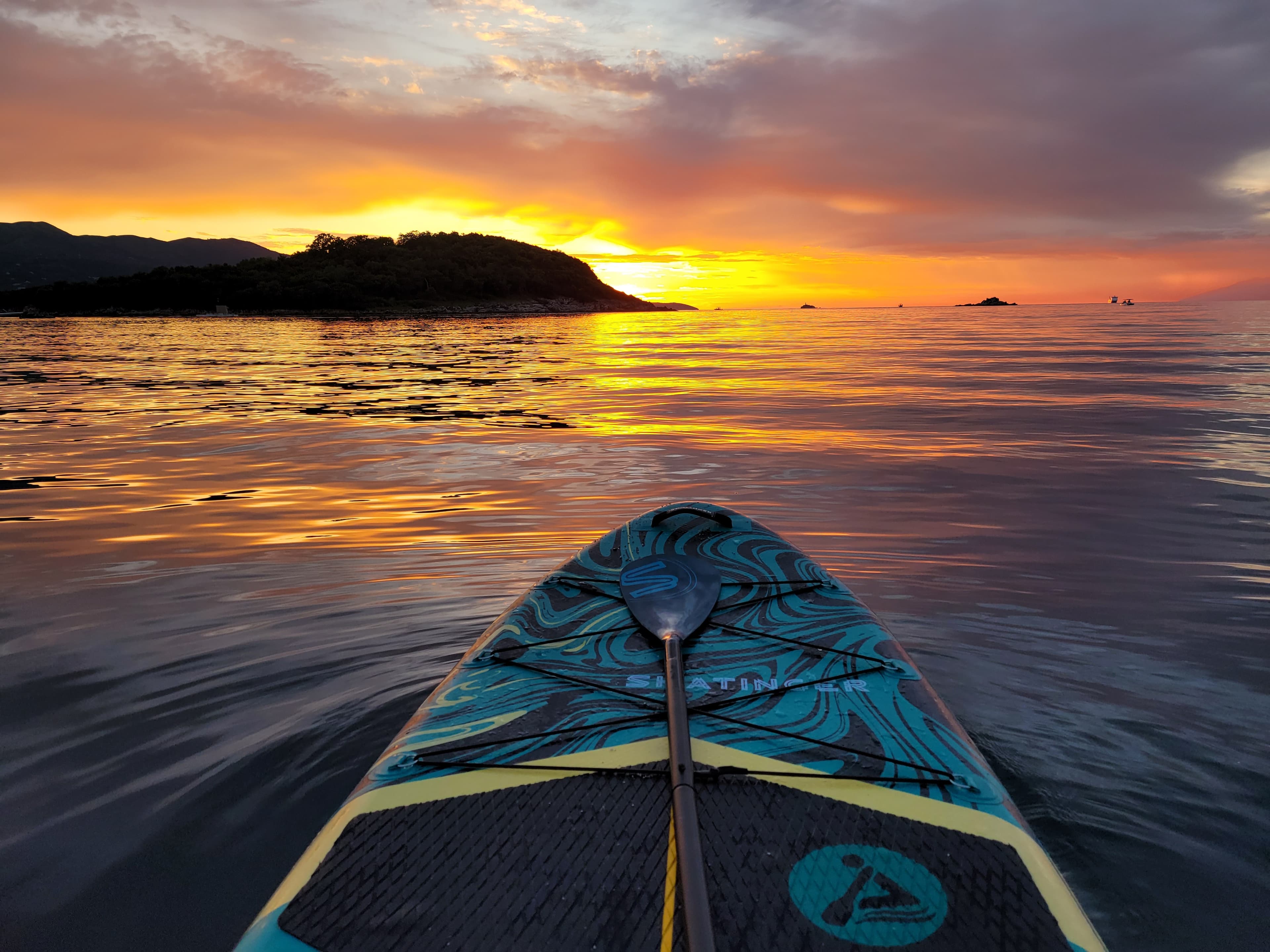 Close-up view from a Stand-Up Paddleboard nose gliding on perfectly smooth, golden reflective water during sunset in Ksamil