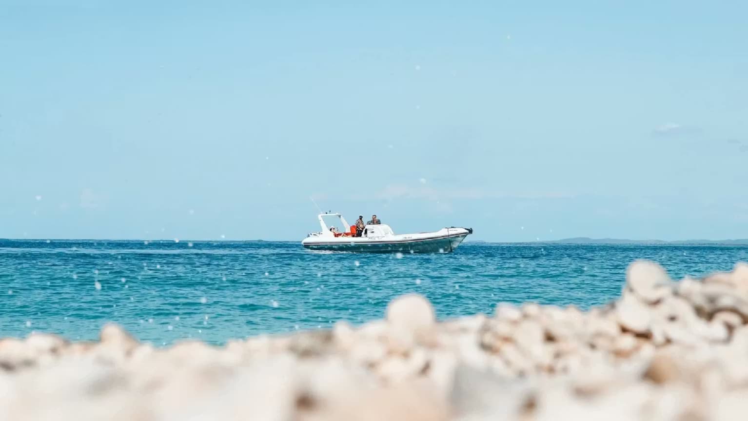View from a white pebble beach of our fast RIB boat cruising through crystal-clear waters on a Ksamil coastal tour